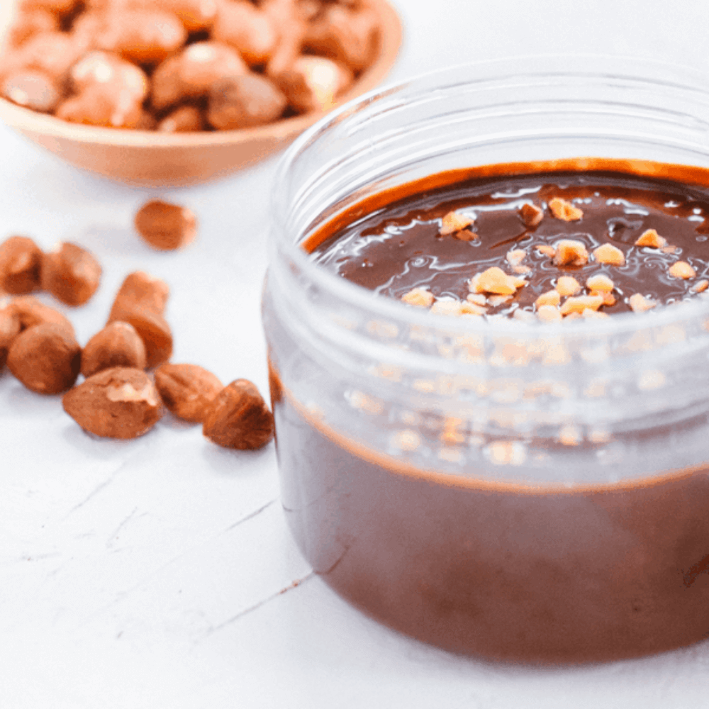 Gezonde keto hazelnoot chocopasta in een glazen pot met hazelnoten op de achtergrond