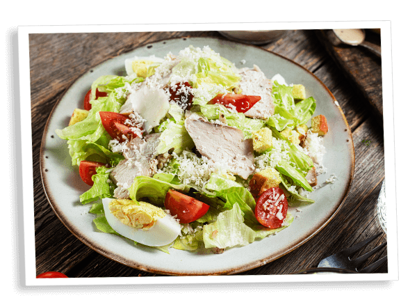 Een Caesar salade op een houten tafel, met knapperige sla, gegrilde kipfilet, gehalveerde cherrytomaatjes, krokante croutons en een hardgekookt ei, afgewerkt met romige dressing en geraspte Parmezaanse kaas — een gezonde, koolhydraatarme maaltijd, ideaal voor een keto- en afslankdieet.