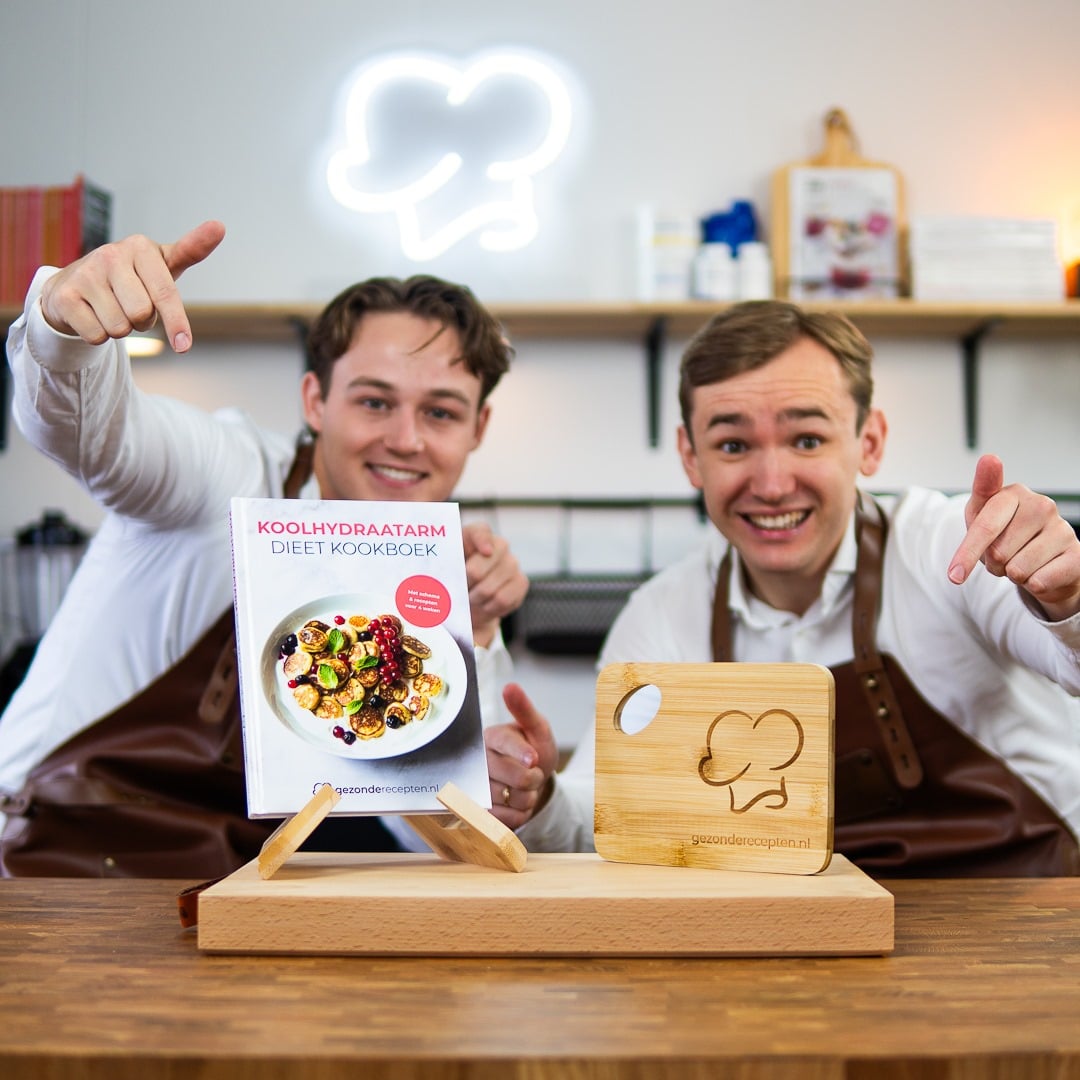 Jay en Mitch, oprichters van Gezonderecepten.nl, presenteren het Koolhydraatarm Dieet Kookboek met mini pannenkoeken, bessen en noten op een romige basis, samen met een houten snijplank met chef-hoed logo en de website "gezonderecepten.nl," ter promotie van een koolhydraatarm dieet.