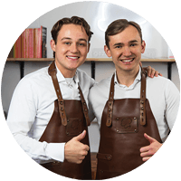 Portret van twee mannen, Jay en Mitch, oprichters van Gezonderecepten.nl, glimlachend en gekleed in witte overhemden met donkerbruine leren schorten, dicht bij elkaar staand met één arm om de ander; keukenplanken met boeken zichtbaar op de achtergrond.