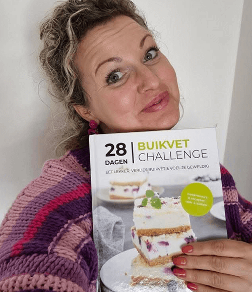 Een vrouw met krullend roodblond haar en bril houdt het Nederlandse kookboek "28 Dagen Buikvet Challenge" van Gezonderecepten.nl vast, met de ondertitel "Heerlijk eten, buikvet verliezen en je geweldig voelen" en een cover met een gelaagd no-bake dessert met romige vulling en rode bessen — een koolhydraatarm dieetboek voor gewichtsverlies