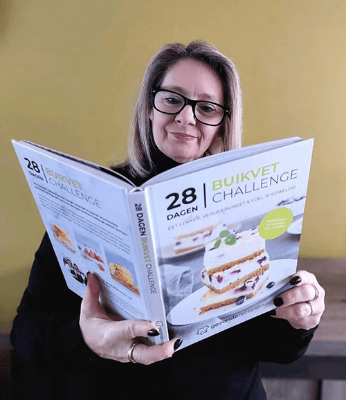 en vrouw met bril en blond haar houdt het hardcover Gezonderecepten.nl kookboek "28 Dagen Buikvet Challenge" vast, een koolhydraatarm dieetboek voor gewichtsverlies met focus op buikvet.