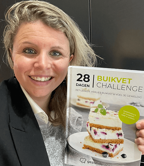 Een vrouw met bril en blond haar houdt het Nederlandse kookboek "28 Dagen Buikvet Challenge" vast, met promotie van koolhydraatarme recepten van Gezonderecepten.nl voor gewichtsverlies.