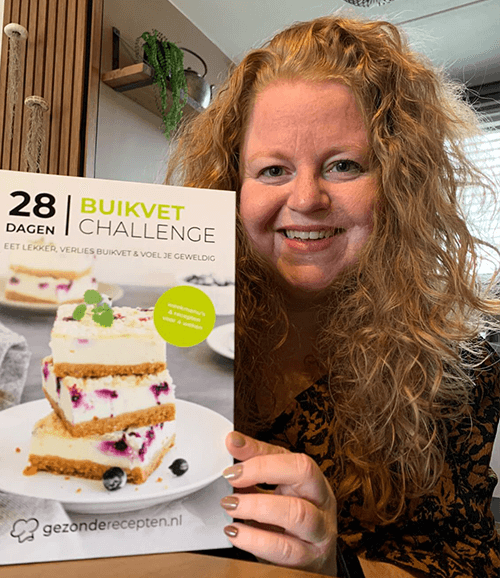 Een vrouw met krullend roodblond haar houdt het Nederlandse kookboek "28 Dagen Buikvet Challenge" van Gezonderecepten.nl vast — een koolhydraatarm dieetboek voor gewichtsverlies.