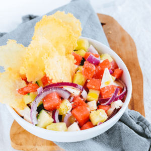 Een witte kom met een verfrissende salade van rode watermeloenblokjes, gele fruitstukjes, feta, gesneden rode ui en komkommer, gegarneerd met grote Parmezaankrullen, geplaatst op een houten plank met grijs textiel eronder — een kleurrijke koolhydraatarme keto maaltijd ideaal voor gezond dieet en gewichtsverlies.