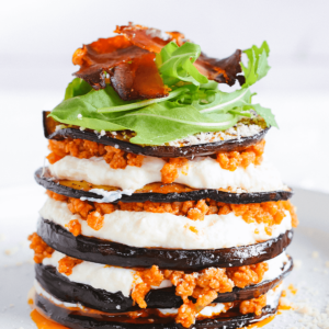 Een hoge, gelaagde keto aubergine Parmezaan schotel met geroosterde aubergine, romige witte saus en vleessaus, afgemaakt met rucola, krokante prosciutto en geraspte kaas — een heerlijke koolhydraatarme maaltijd voor gezond dieet en gewichtsverlies.