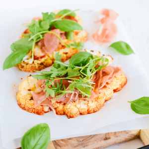 Twee geroosterde bloemkoolsteaks met prosciutto, rucola en verse basilicumblaadjes op bakpapier — een smaakvol koolhydraatarm en keto-vriendelijk gerecht voor gezond eten en gewichtsverlies.
