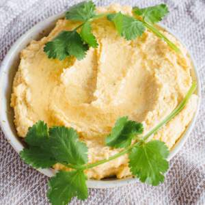 Een kom met romige, lichtgele hummus met een scheutje olijfolie in het midden en gegarneerd met verse koriander, geplaatst op een lichtgekleurde, getextureerde doek — een zachte koolhydraatarme keto dip ideaal voor gezond dieet en gewichtsverlies.