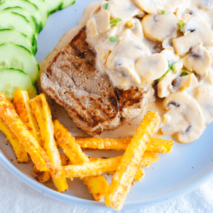 Een wit bord met een sappige biefstuk overgoten met romige champignonsaus en kruiden, geserveerd met schijfjes komkommer aan de zijkant en een portie goudbruine friet op de voorgrond — een stevige koolhydraatarme keto maaltijd ideaal voor gezond dieet en gewichtsverlies.