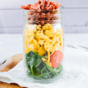 Een doorzichtige glazen mason jar met lagen van verse spinazie, verkruimelde gele kaas, gele cherrytomaten, luchtige roerei en verkruimeld spek of worst bovenop — een handige koolhydraatarme keto maaltijd voor ontbijt of lunch, ideaal voor gezond dieet en gewichtsverlies.