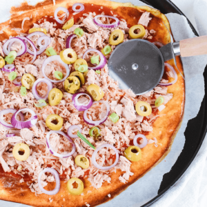 Een dunne pizza met tonijnvlokken, groene olijven en rode uiringen op een donkere ronde bakplaat met een pizzasnijder ernaast, een gezond koolhydraatarm en keto-vriendelijk gerecht