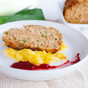 Een stevige plak keto gehaktbrood met stukjes ui en kruiden, geserveerd op een wit bord met gebakken gele uien en een rode bessensaus, terwijl in de achtergrond de ovenschaal met de rest van het gehaktbrood te zien is — een smaakvolle koolhydraatarme maaltijd voor gezond dieet en gewichtsverlies.