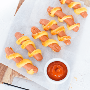Meerdere hotdogs gewikkeld in goudbruin deeg, netjes gerangschikt op bakpapier op een houten plank, met een klein wit kommetje rode ketchup als dipsaus — een leuke koolhydraatarme keto snack ideaal voor gezond dieet en gewichtsverlies.