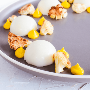 Een verfijnd bloemkoolgerecht met drie witte domes, geroosterde en rauwe bloemkoolroosjes en gele puree, gepresenteerd in moderne fine-dining stijl, ideaal voor een koolhydraatarm of keto dieet