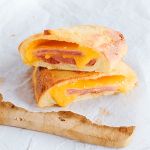 Twee helften van een goudbruin gebakken broodje gevuld met plakjes ham en gesmolten oranje kaas, waarbij de kaas tussen de lagen knapperig deeg uitloopt en een stukje deeg op de voorgrond ligt — een heerlijke koolhydraatarme keto snack ideaal voor gezond dieet en gewichtsverlies.