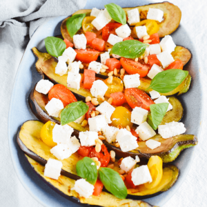 Geroosterde aubergineplakken op een lichtblauwe schaal, gegarneerd met rode en gele cherrytomaten, verkruimelde feta, pijnboompitten en verse basilicum — een Mediterraan koolhydraatarm keto salade- of bijgerecht ideaal voor gezond dieet en gewichtsverlies.
