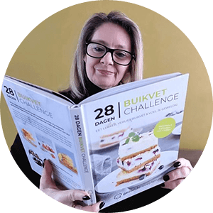 Een vrouw met bril en blond haar houdt het hardcover kookboek "28 Dagen Buikvet Challenge" van Gezonderecepten.nl vast, met op de cover een gelaagd no-bake dessert met romige vulling en rode bessen — een koolhydraatarm dieetboek voor gewichtsverlies met focus op buikvet.