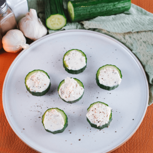 Zes keto komkommerhapjes met een romige kaasvulling, bestrooid met zwarte peper en kruiden, geserveerd op een licht bord met knoflook en komkommers op de achtergrond — een frisse, low-carb snack of voorgerecht.