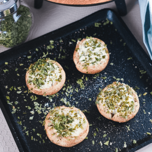 Zwart rechthoekig bakblik met vier gevulde champignons met romige witte vulling en gehakte groene kruiden, met een pot gedroogde kruiden en een blauw-wit servet op de achtergrond, ideaal voor keto, low-carb en gewichtsverlies