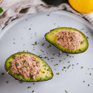Twee avocadohelften gevuld met tonijnsalade, gegarneerd met gesneden bieslook en zwarte peper, geserveerd op een grijze schaal met een citroen en gestreepte keukendoek op de achtergrond, ideaal voor keto, low-carb en gezond eten