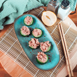 Tonijnsalade geserveerd op komkommerschijfjes, gegarneerd met rode pepervlokken op een bord, met een halve citroen, kruidenpotjes en eetstokjes op de achtergrond, ideaal voor keto en lowcarb diëten.
