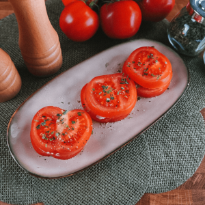 Ovaal bord met tomaatplakjes, gekruid met kruiden en zwarte peper, met hele tomaten en kruidenmolens op de achtergrond, ideaal voor afvallen en keto dieet