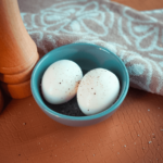 Twee gepelde gekookte eieren bestrooid met zwarte peper in een klein blauw kommetje, een keto- en low-carb snack voor gezond afvallen, op een bruine textuur ondergrond met een handdoek en pepermolen ernaast