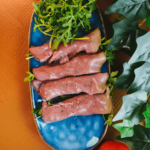 Keto en low-carb dun gesneden rosbief geserveerd op verse rucola voor gezond gewichtsverlies, gekruid met zwarte peper, met een rode tomaat en groene bladeren op een bruine ondergrond