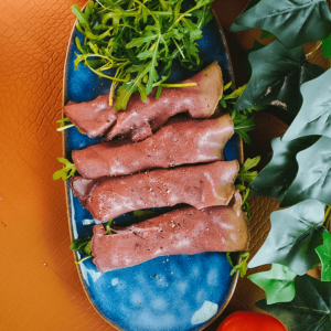 Keto en low-carb dun gesneden rosbief geserveerd op verse rucola voor gezond gewichtsverlies, gekruid met zwarte peper, met een rode tomaat en groene bladeren op een bruine ondergrond