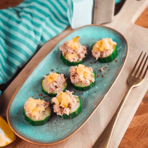 Zes komkommerschuitjes gevuld met tonijnsalade en gegarneerd met stukjes ananas op een teal-ovaal bord, bestrooid met zwarte peper en kruiden, geserveerd met een schijfje citroen en een vork op een houten snijplank, ideaal voor keto- en low-carb diëten