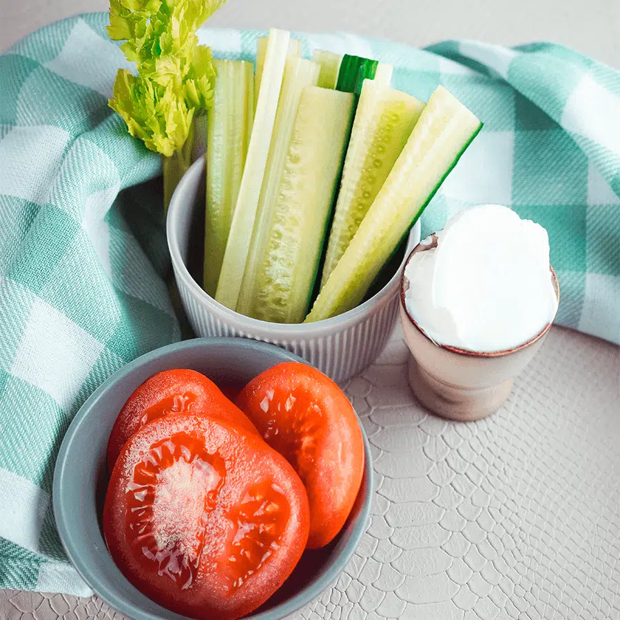Een groenteschotel met komkommer- en selderijstengels in een witte kom, gehalveerde rode tomaten in een grijze kom en een kleine kom met romige yoghurtdip, allemaal gerangschikt op een grijs oppervlak met een groen-wit geruit keukendoekje, ideaal voor gezonde keto- en low-carb diëten