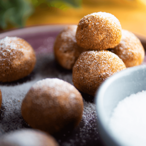 Koolhydraatarme en eiwitrijke donutballetjes bestrooid met poedersuiker, geserveerd op een donkerpaars bord met een klein kommetje suiker erbij. Perfect voor keto en gezond dieet.