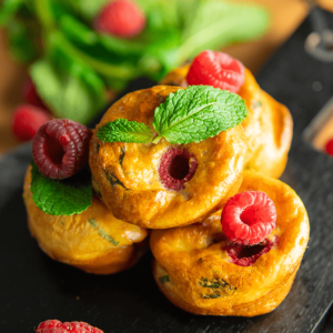 Goudbruine keto-muffins genaamd Raspberries-Mascarpone-Cloud-Cakejes, getopt met verse frambozen en munt, met een vochtige, luchtige textuur en zichtbare stukjes framboos, geplaatst op een donkere, getextureerde ondergrond met extra frambozen en munt eromheen.