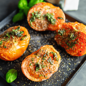 Vier halve geroosterde tomaten met kruiden, specerijen en kaas op een zwart bord, ideaal voor keto, low-carb en gezond dieet.