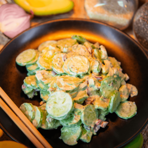 Een zwart bord met romige keto komkommersalade van dun gesneden komkommer en rode ui, bedekt met een pittige dressing, met houten eetstokjes aan de zijkant.