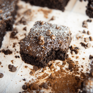 Een fudgy vierkante keto brownie, getopt met chocoladechips en poedersuiker, op een witte ondergrond met verspreide chocoladechips en cacaopoeder.