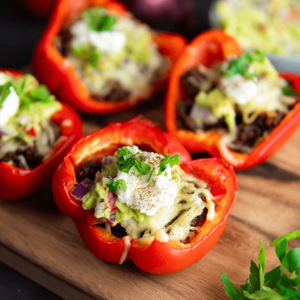Meerdere rode paprika’s doormidden gesneden en gevuld met een keto-vriendelijk gehaktmengsel, getopt met romige saus, avocado-topping en gegarneerd met gehakte kruiden en zwarte peper op een houten snijplank.