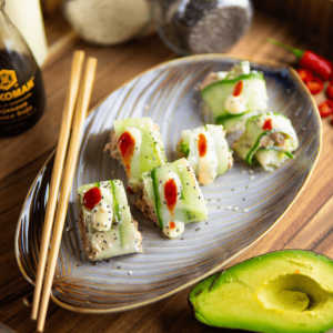 Kleine komkommer sushi rolls gevuld met tonijnsalade, gegarneerd met romige en pittige saus en zwarte sesamzaadjes, geserveerd met avocado, ideaal voor keto en low-carb dieet.