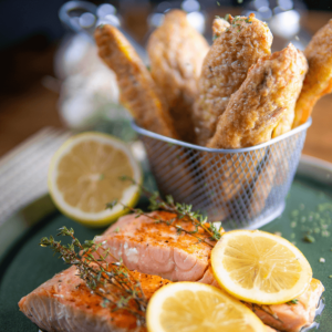 Low-carb fish and chips met twee stukken gebakken zalm, gegarneerd met verse tijm en citroenschijfjes, geserveerd op een donkergroen bord met krokante vissticks op de achtergrond, ideaal voor keto en gezond dieet.