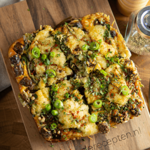 Vierkante pizza met dikke korst, belegd met gesmolten kaas, champignons, olijven, groene ui, kruiden en rode pepervlokken op een houten plank, keto- en low-carb recept van Gezonderecepten.nl.