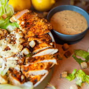 Een keto-vriendelijke kip Caesar salade met gegrilde kipfilet, romaine sla, croutons en een romige dressing in een klein kommetje, geserveerd met citroenpartjes op de achtergrond.
