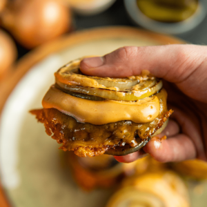 Een hand die een Flying Dutchman burger vasthoudt zonder broodje, met sappige runderburgers, gesmolten kaas en gegrilde uien — een ideale koolhydraatarme maaltijd voor gewichtsverlies en een gezond dieet.