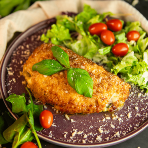 Een bord met krokante gepaneerde kip Milanese, gegarneerd met basilicum en Parmezaanse kaas, geserveerd met een salade van gemengde groenten en cherrytomaten — een eiwitrijke, koolhydraatarme keto maaltijd, ideaal voor gewichtsverlies en een gezond dieet