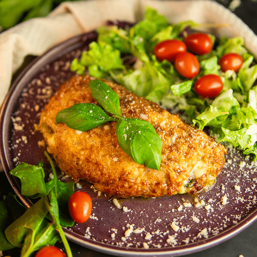 Een bord met krokante gepaneerde kip Milanese, gegarneerd met basilicum en Parmezaanse kaas, geserveerd met een salade van gemengde groenten en cherrytomaten — een eiwitrijke, koolhydraatarme keto maaltijd, ideaal voor gewichtsverlies en een gezond dieet