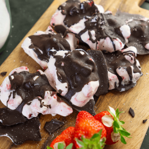 Een batch koolhydraatarme aardbeien yoghurt clusters bedekt met pure chocolade, gepresenteerd op een houten snijplank met verse aardbeien — een perfect tussendoortje voor een koolhydraatarm dieet