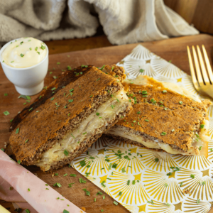 Een gegrilde koolhydraatarme toast doorgesneden, met gesmolten kaas binnenin, geserveerd met een romige dipsaus met bieslook en plakjes ham op de achtergrond — een koolhydraatarme maaltijd voor een gezond dieet