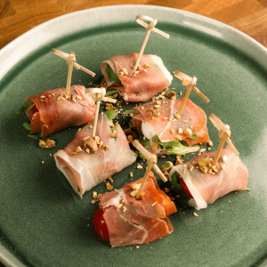 Een groen bord met Keto rolletjes van prosciutto gevuld met kaas, rucola en stukjes tomaat of meloen, gegarneerd met gehakte noten. Een koolhydraatarme snack, ideaal voor gewichtsverlies en een gezond dieet.