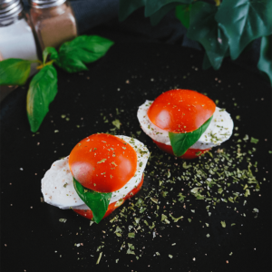 Een donker bord met gestapelde Caprese salade hapjes van verse mozzarella, gehalveerde cherrytomaten en basilicumblaadjes, gegarneerd met kruiden — een keto en koolhydraatarm voorgerecht voor gezond eten en gewichtsverlies