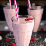 Drie hoge glazen met romige frambozen milkshakes, gegarneerd met verse frambozen en gestreepte rietjes, geserveerd op een donker aanrecht, een gezonde en verfrissende drank voor een gezond dieet