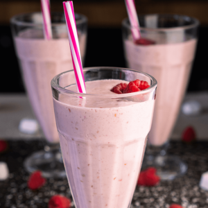 Drie hoge glazen met romige frambozen milkshakes, gegarneerd met verse frambozen en gestreepte rietjes, geserveerd op een donker aanrecht, een gezonde en verfrissende drank voor een gezond dieet